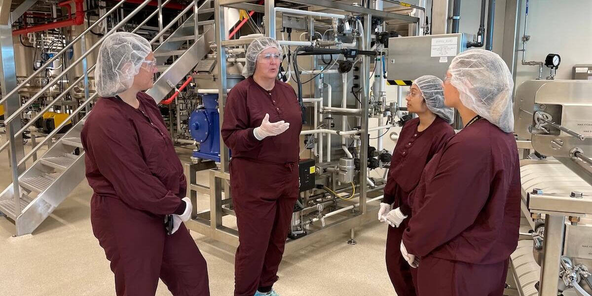 Two individuals participating in the Maryland Chamber Foundation's Teacher Externship Program stand in a group while wearing scrubs and protective gear with factory equipment behind them.